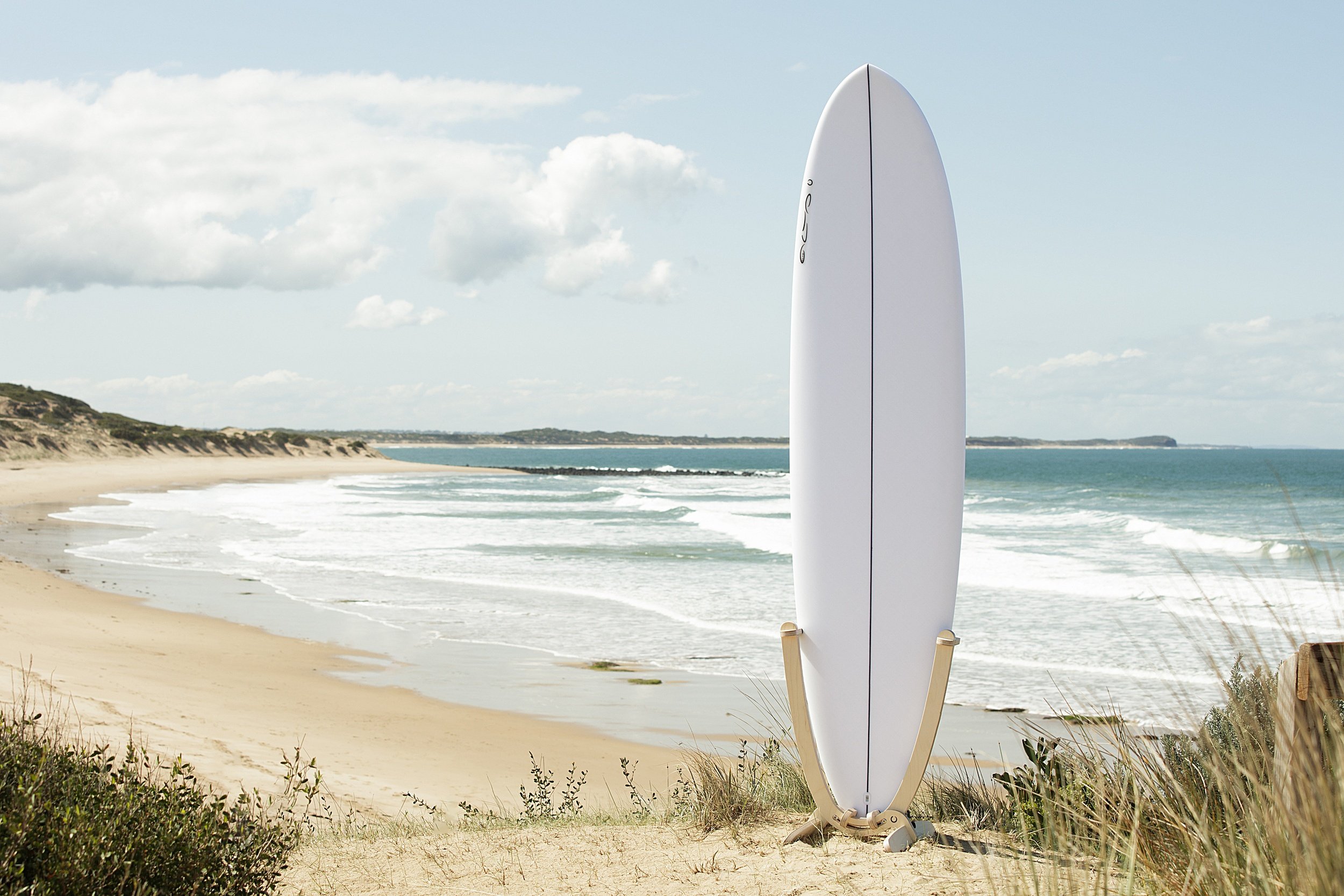 An all white Surfboard with a GLYD O decal on its nose, is mounted in a GLYD O surfboard rack. The rack with board is standing on a sandy dune with the beach, waves and ocean in the back ground, coastal dunes in the distance.
The Surfboard Rack is a Beautiful birchwood curved single surfboard rack in the shape of the tail of a surfboard, with hooks to hold the rails and tail and two slightly curved feet connected perpendicular to the upright curved arms. Image is taken from a slight angle in relations to the vertical surfboard rack. Rectangular image.